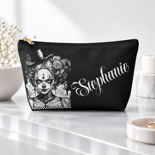 A black cosmetic pouch with a gold zipper, featuring a black and white graphic of a scary clown girl with roses and the name 'Stephanie' in white script, displayed on a reflective white surface. A personalizable bag.